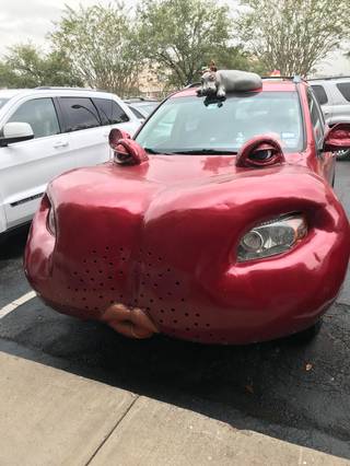 A red car in a parking lot has large sculpted eyes on the hood and lips on the front bumper.