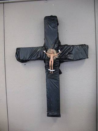 A doll-like figure is attached to a dark cross-shaped object.