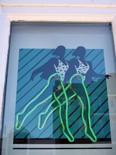 Two green neon hangers aligned over painted swimsuit figures in a shop window.