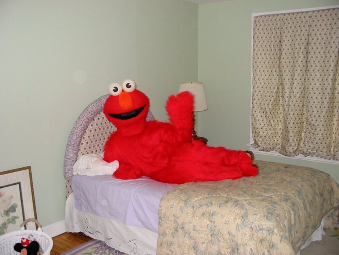 A bright red Elmo plush lies alone on a made bed.