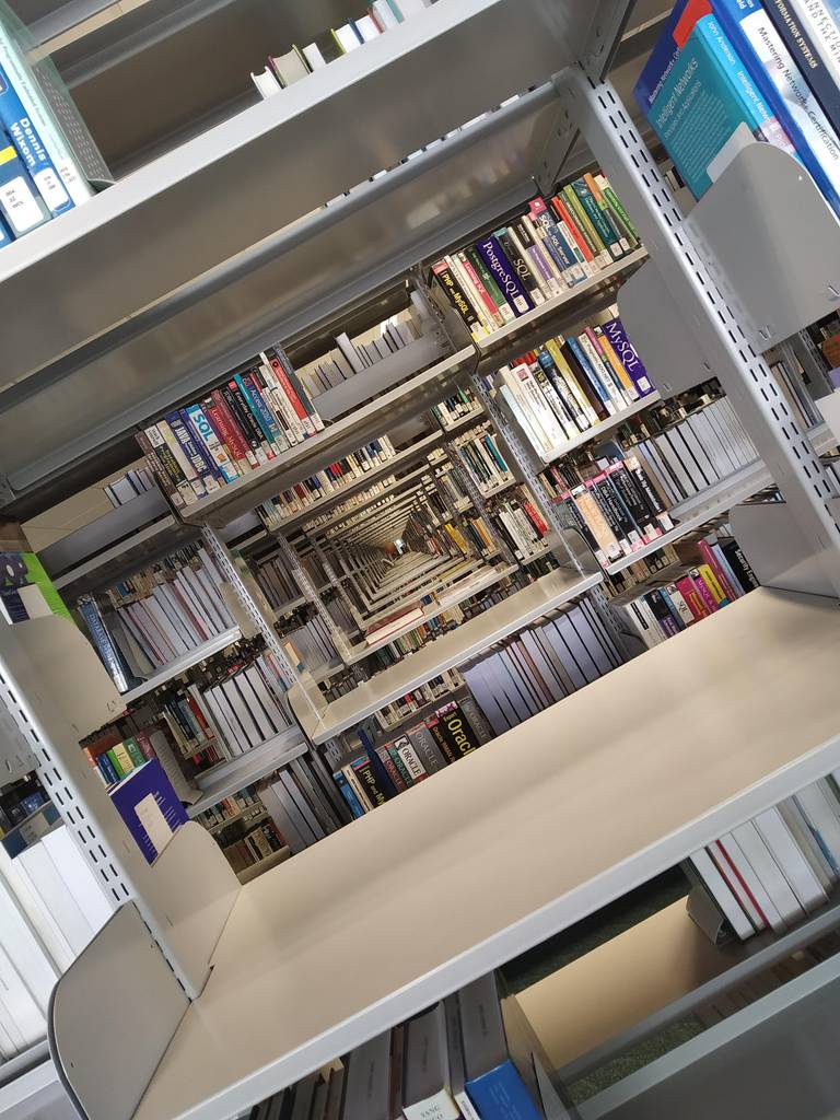 A library aisle photographed at a rotated angle, with bookshelves extending across the frame like walls.