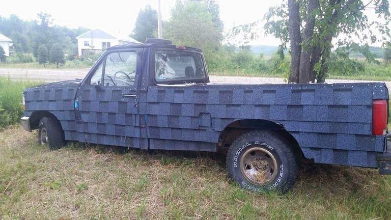 A parked pickup truck has blue asphalt roofing shingles attached across its hood, doors, and bed.