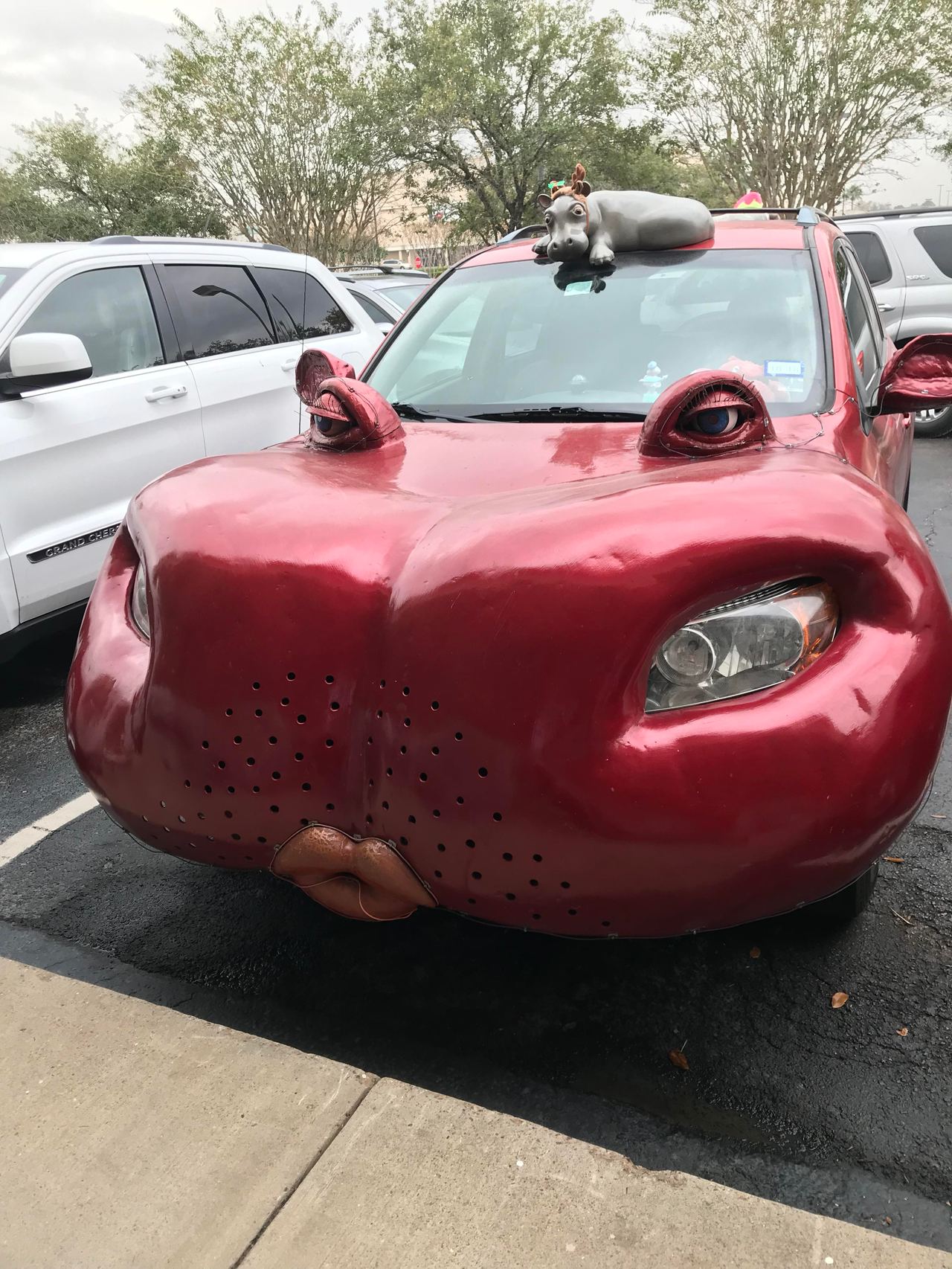 A red car in a parking lot has large sculpted eyes on the hood and lips on the front bumper.