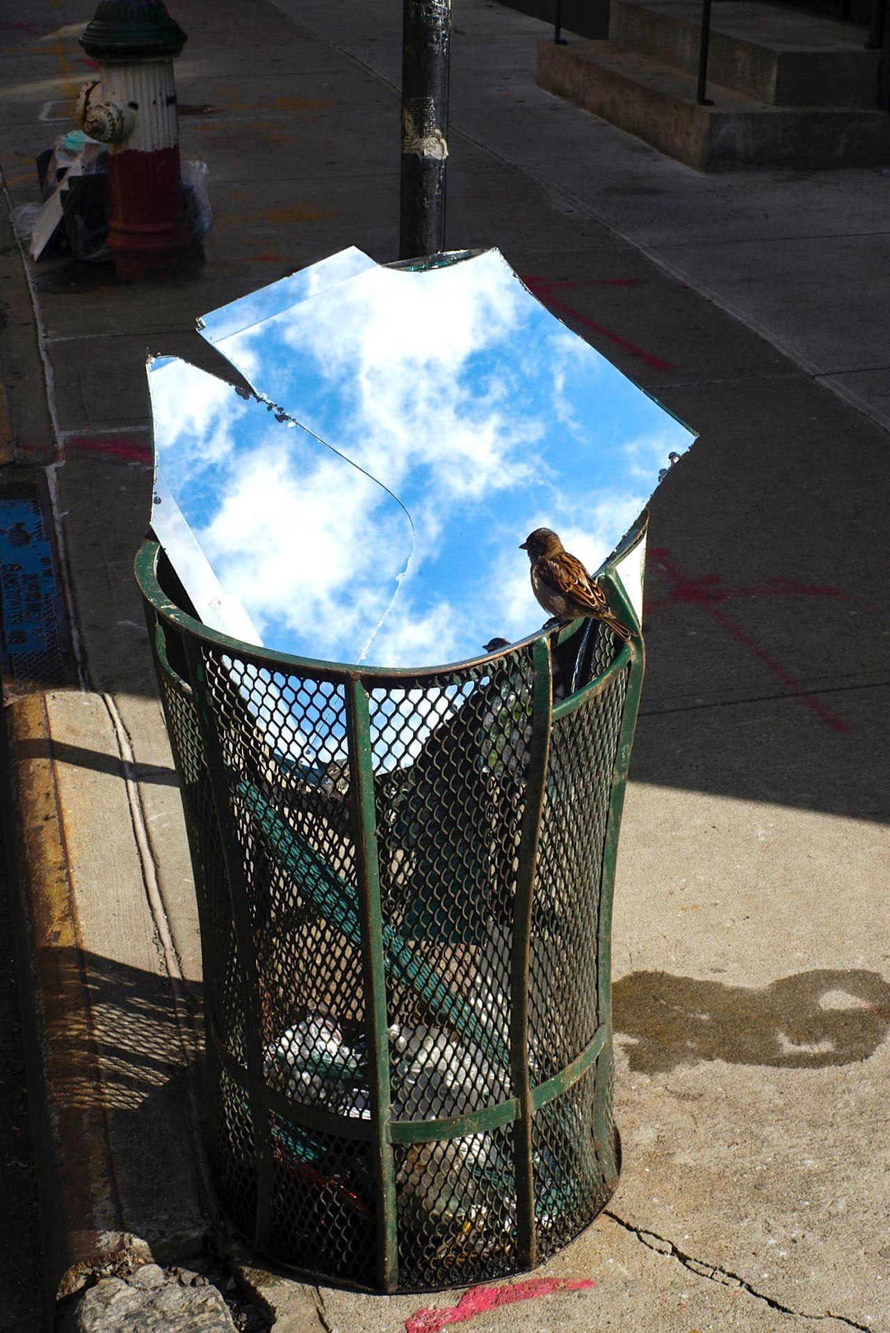 Mirror panels rest over a metal street bin and reflect blue sky and white clouds.