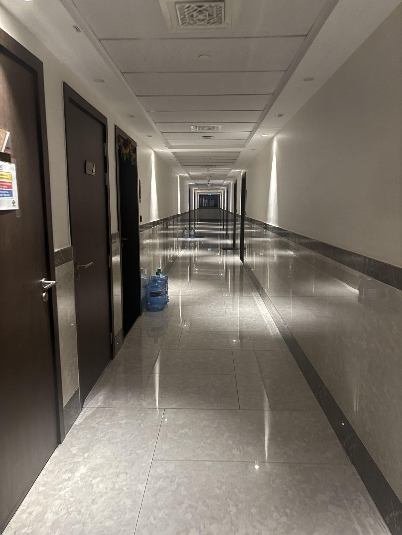 A long empty hallway with dark doors, glossy tiles, reflective wall panels, and water bottles.