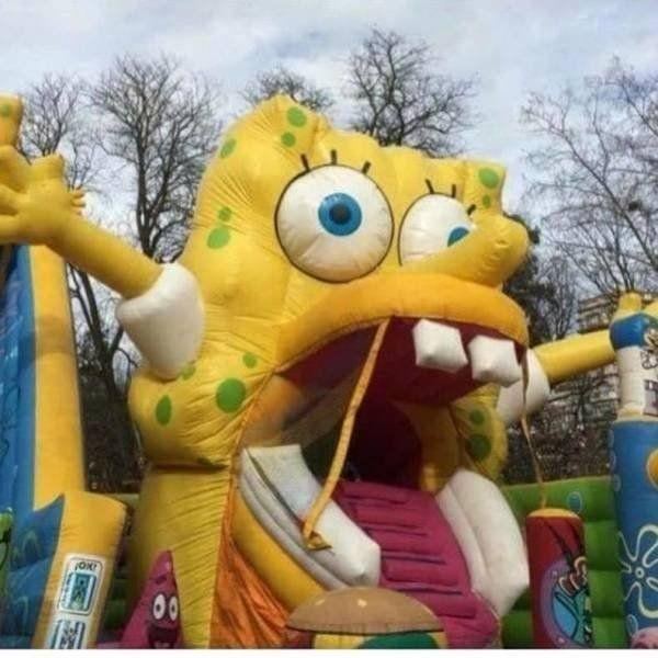 A yellow square character inflatable stands outdoors with mismatched eyes, large teeth, and a wide open mouth entrance.