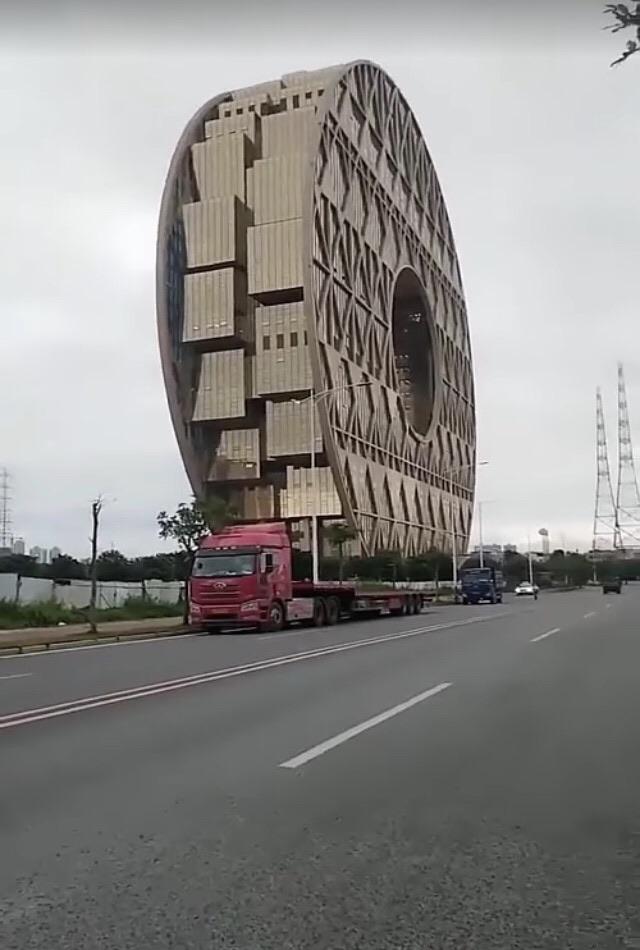 A flatbed truck parked in front of an oval building appears to be carrying the building.