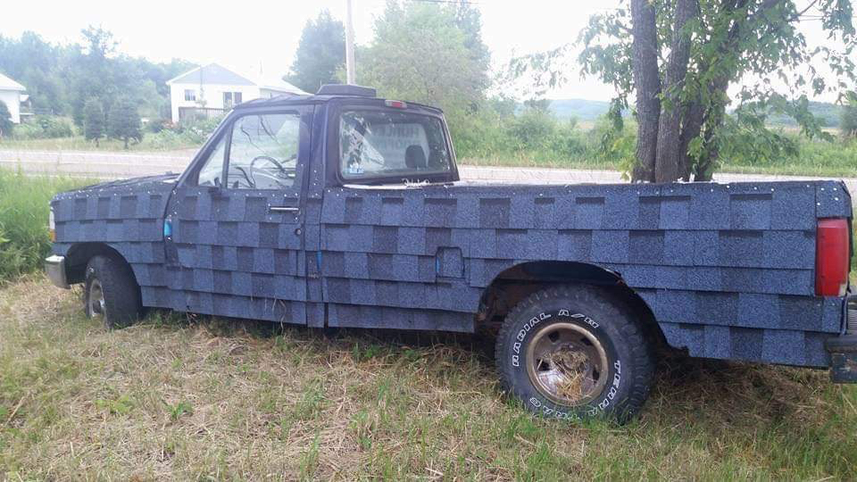 A parked pickup truck has blue asphalt roofing shingles attached across its hood, doors, and bed.