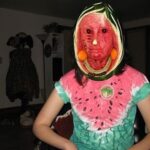 Cursed image of a human with a watermelon mask in a dimly lit room.