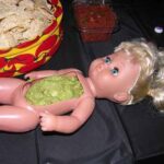 A doll's stomach hollowed out and filled with guacamole, surrounded by chips.