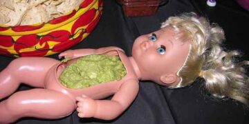 A doll's stomach hollowed out and filled with guacamole, surrounded by chips.