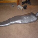 A human-like figure completely wrapped in duct tape lying on a carpeted floor.