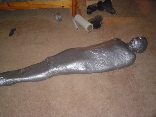 A human-like figure completely wrapped in duct tape lying on a carpeted floor.
