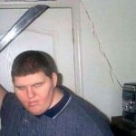 A machete-wielding man posing in front of a door in a bedroom.