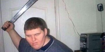 A machete-wielding man posing in front of a door in a bedroom.