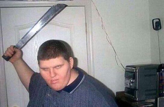 A machete-wielding man posing in front of a door in a bedroom.